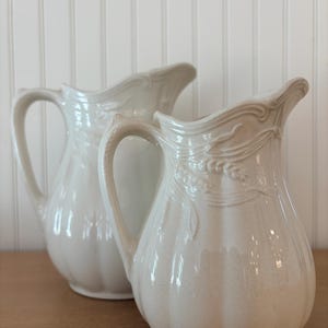 Puede incluir: Dos jarras de cerámica blanquecina con asas curvas y relieves florales decorativos. Las jarras están sobre una superficie de madera contra una pared blanca con paneles. Una jarra tiene una superficie lisa, la otra es texturizada.
