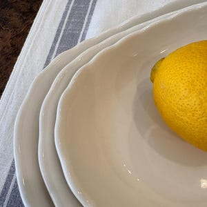 May include: A stack of three off-white, scalloped-edge ceramic plates. A bright yellow lemon rests on the top plate. The plates are on a white and blue striped cloth, suggesting a kitchen or dining setting.
