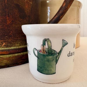May include: A white ceramic mug featuring a green watering can design with a rabbit peeking out. The mug has text on the side. Other rustic pottery is in the background.