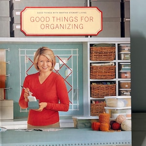 Puede incluir: Portada de libro con el título "Good Things for Organizing" de Martha Stewart. La imagen presenta a una mujer con un suéter rojo sosteniendo una pequeña caja, con estantes de cestas y recipientes transparentes en el fondo. El tema general es la organización del hogar.