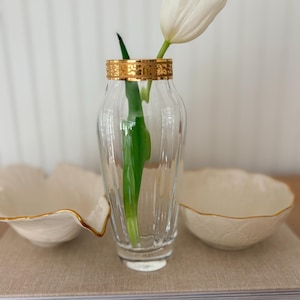 May include: A clear glass vase with a gold rim holds a white tulip. Two small, off-white decorative bowls with gold trim are on either side of the vase. The items are arranged on a neutral-toned surface.