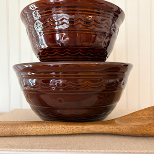 May include: Two stacked, glossy brown ceramic mixing bowls with a decorative wave pattern around the rim. A wooden spoon rests on a stack of books in the foreground. The bowls are a rich, warm brown color.