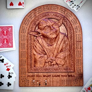 May include: A wooden cribbage board with a carved image of Yoda from Star Wars. The board has a brown stain and the text "PLAY YOU MUST. LOSE YOU WILL." is engraved below the image.