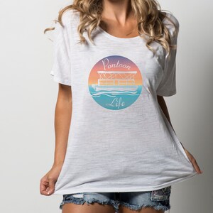 Pu&ograve; includere: Maglietta bianca oversize con una grafica di una barca ponton su uno sfondo di tramonto. La grafica dice "Pontoon Life".