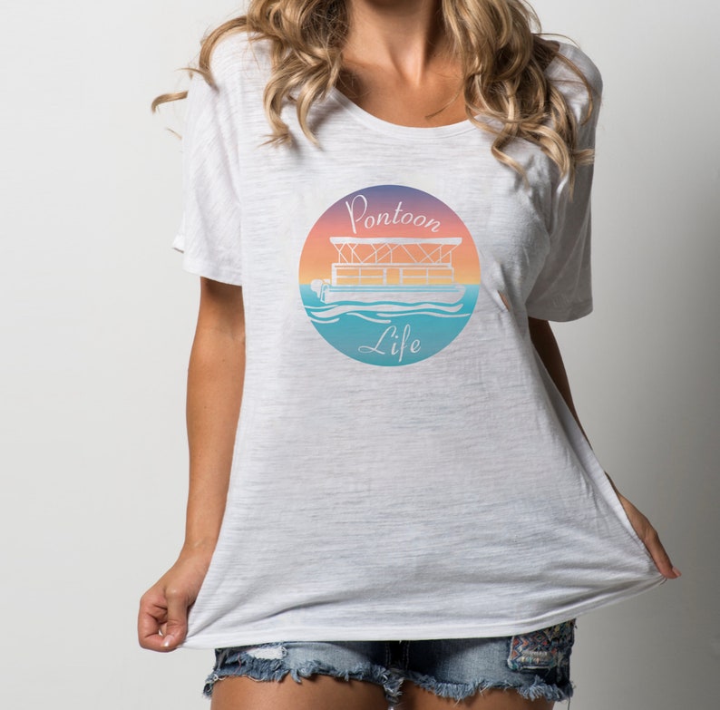 Pu&ograve; includere: Maglietta bianca oversize con una grafica di una barca ponton su uno sfondo di tramonto. La grafica dice "Pontoon Life".