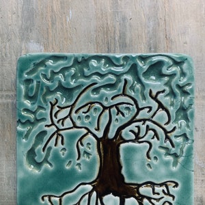 May include: A square ceramic tile with a raised design of a tree with roots and branches. The tile is a light blue-green color with brown accents.