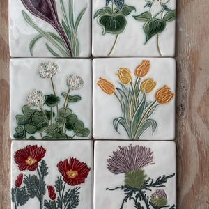 May include: A set of six square ceramic tiles, each featuring a raised, hand-painted botanical illustration. The designs include a purple crocus, white trilliums, white clover, yellow tulips, red poppies, and a purple thistle, all on white backgrounds.