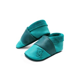 Könnte beinhalten: Ein Paar babyblaue Lederschuhe mit Teddybär-Motiv. Die Schuhe haben ein weiches Wildlederfutter und eine flexible Sohle.
