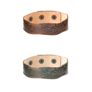 Pode incluir: Duas pulseiras de couro com um design floral. A pulseira superior é marrom com um fundo marrom claro. A pulseira inferior é marrom escuro com um fundo marrom claro.