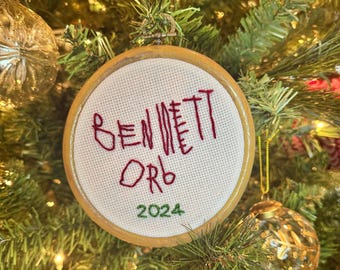 May include: A round, embroidered Christmas ornament with the name "BENNETT ORB" in red thread. The year "2024" is stitched in green. The ornament is set in a wooden hoop and hangs on a Christmas tree.