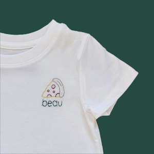 May include: White t-shirt with a small embroidered pizza slice design. The pizza slice has yellow, red, and brown details. Below the pizza is the word "beau" embroidered in green thread. The shirt has short sleeves and a crew neck.