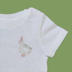 May include: A white short-sleeved t-shirt featuring an embroidered design on the chest. The embroidery depicts a white goose wearing a small red party hat, with the name XAVIER stitched in black thread next to the goose.