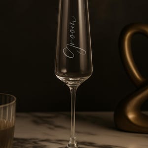 May include: A clear champagne flute with the word "Groom" elegantly scripted on the glass. The slender stem and wide base provide stability. The glass is set against a dark background with a gold decorative element.