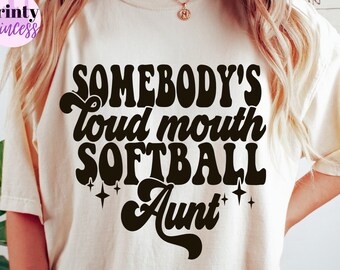 May include: A white t-shirt with black text that reads "Somebody's loud mouth softball aunt" in a retro font. The text is surrounded by small stars.