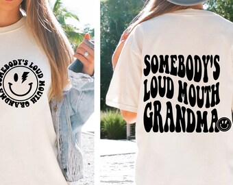 May include: White t-shirt with a black graphic that says "Somebody's Loud Mouth Grandma" with a smiley face. The graphic is in a retro font.