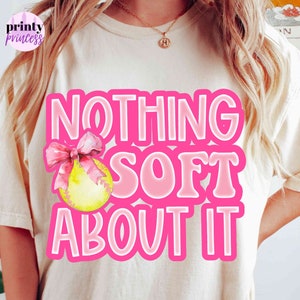 May include: A white t-shirt with a pink graphic that says "NOTHING SOFT ABOUT IT" with a pink ribbon and a yellow softball.