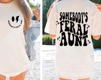 May include: White t-shirt with a black graphic that says "Somebody's Feral Aunt" in a retro font with a smiley face and lightning bolt above the text.