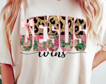 May include: Cream-colored t-shirt featuring the words "JESUS wins" in a bold, decorative font. The letters have a leopard print and floral design, with a pink bow. The text is in black script.