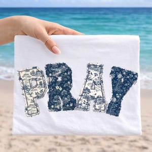 May include: White t-shirt with the word "PRAY" in large, decorative letters. The letters are filled with a blue and white toile pattern featuring landscapes and floral designs. The shirt is held up against a beach and ocean backdrop.