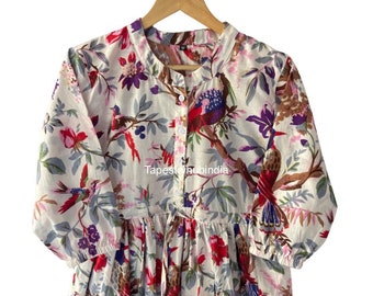 May include: A white cotton dress with a floral print featuring birds and flowers. The dress has a button-up front and a gathered skirt. The dress is hanging on a wooden hanger.