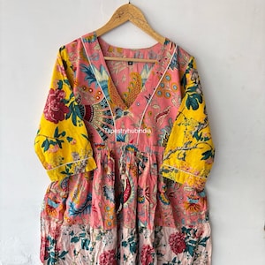 May include: A pink, yellow and red floral print cotton dress with a V-neckline and 3/4 sleeves. The dress has a patchwork design and a white trim around the neckline. The dress is hanging on a wooden hanger. "Tapestryhubindia" is printed on the dress.