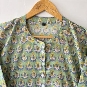 May include: A green cotton shirt with a yellow and blue floral print. The shirt has a mandarin collar and a button closure. The shirt is hanging on a wooden hanger.