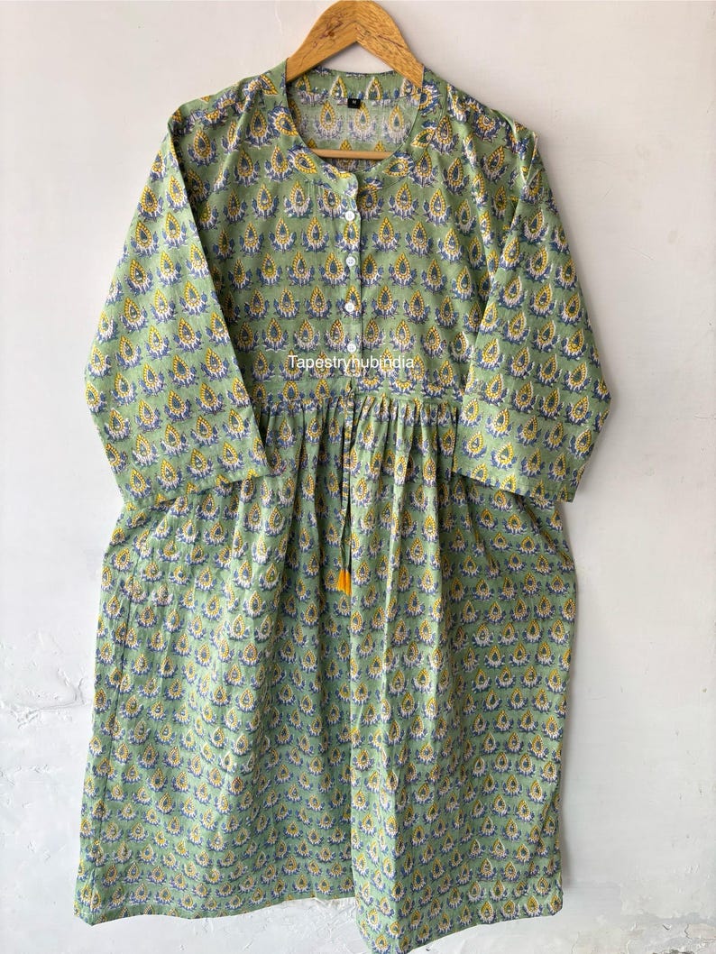 May include: A green cotton dress with a yellow and blue floral print. The dress has a round neckline, three-quarter sleeves, and a drawstring waist with a tassel. The dress is made in India and is available in sizes S-XL.