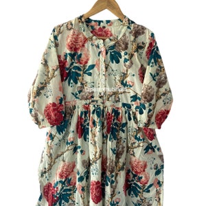 May include: A white cotton dress with a floral print featuring large pink and red flowers, green leaves, and brown branches. The dress has a button-up front and a gathered waist.
