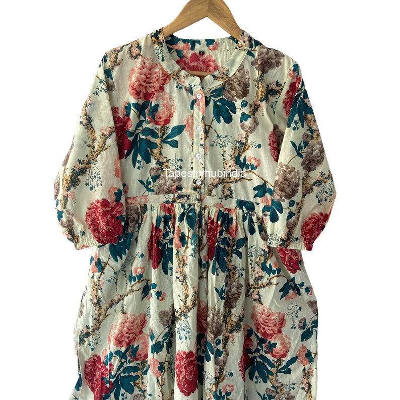May include: A white cotton dress with a floral print featuring large pink and red flowers, green leaves, and brown branches. The dress has a button-up front and a gathered waist.