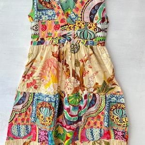 Op de afbeelding: Een mouwloze, veelkleurige patchworkjurk met een V-hals en een strik in de taille. De jurk heeft een bloemenprint en een patchworkontwerp in verschillende kleuren, waaronder rood, roze, blauw, groen en geel.
