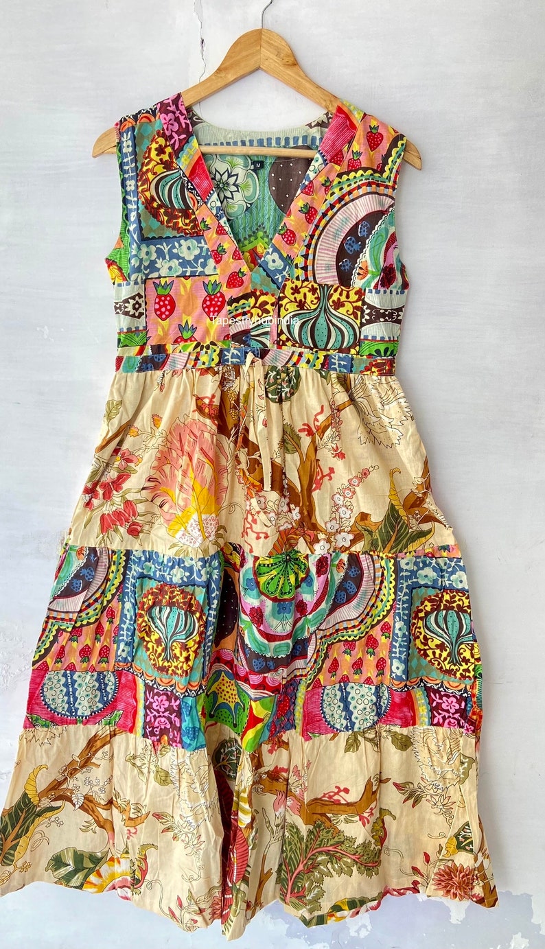 Op de afbeelding: Een mouwloze, veelkleurige patchworkjurk met een V-hals en een strik in de taille. De jurk heeft een bloemenprint en een patchworkontwerp in verschillende kleuren, waaronder rood, roze, blauw, groen en geel.