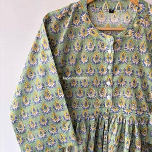 May include: A green cotton tunic with a button-up front and a floral print. The print features yellow, blue, and white teardrop shapes.