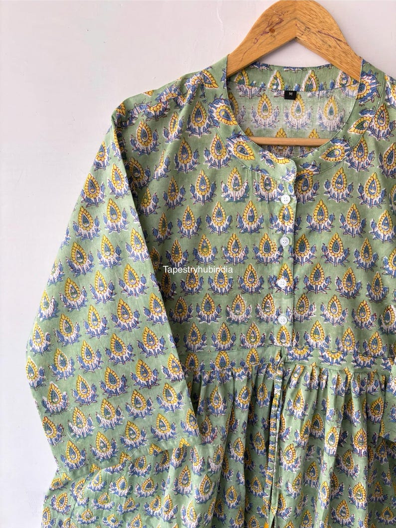 May include: A green cotton tunic with a button-up front and a floral print. The print features yellow, blue, and white teardrop shapes.