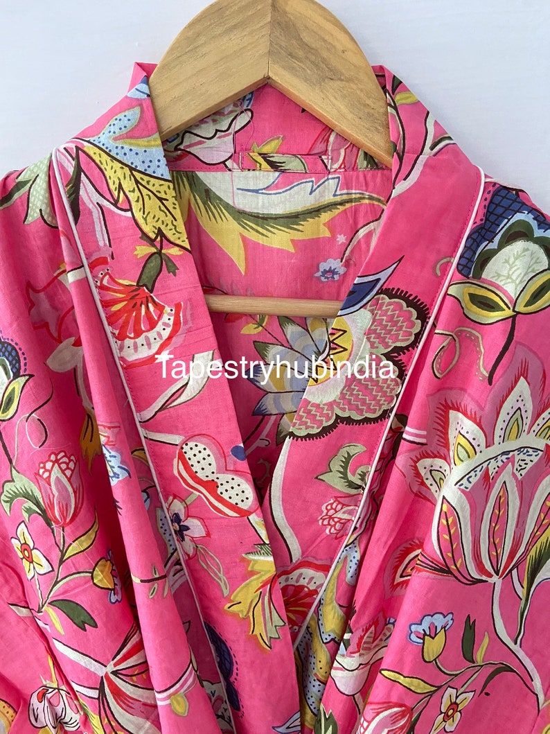 May include: Pink cotton robe with a floral print and white piping. The robe has a shawl collar and a tie closure. The fabric is soft and comfortable.