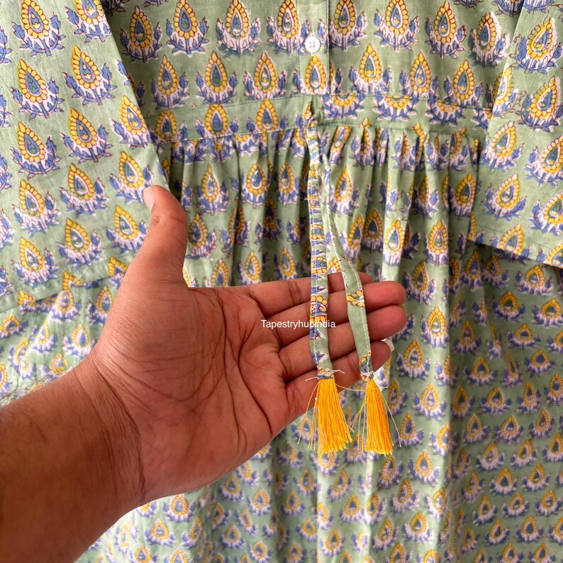 May include: A green cotton dress with a floral print and a drawstring waist. The dress has a button closure at the neck and short sleeves. The drawstring has yellow tassels.