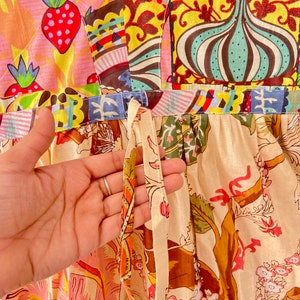 Op de afbeelding: Een witte katoenen jurk met een patchwork-ontwerp met aardbeien, bloemenpatronen en geometrische vormen. De jurk heeft een strik in de taille en een cr&egrave;mekleurige bloemenprint op de rok.