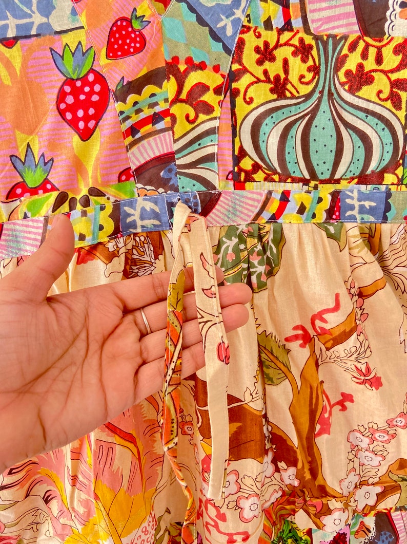 Op de afbeelding: Een witte katoenen jurk met een patchwork-ontwerp met aardbeien, bloemenpatronen en geometrische vormen. De jurk heeft een strik in de taille en een cr&egrave;mekleurige bloemenprint op de rok.