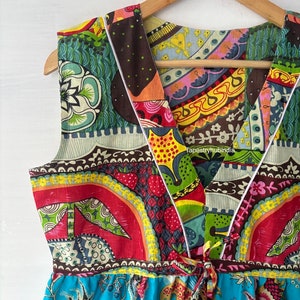 May include: A colourful, patchwork-style dress with a V-neckline and a tie at the waist. The dress is made of a variety of fabrics with different patterns, including floral, geometric, and paisley. The dress is sleeveless and has a flowing skirt.