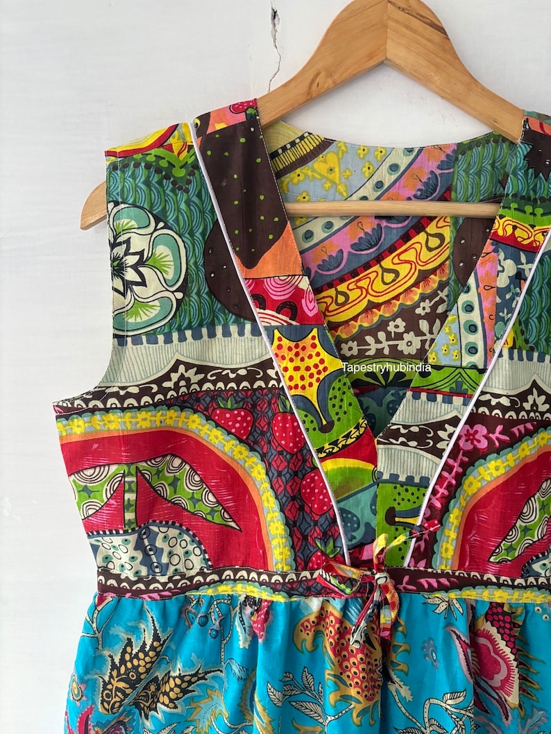 May include: A colourful, patchwork-style dress with a V-neckline and a tie at the waist. The dress is made of a variety of fabrics with different patterns, including floral, geometric, and paisley. The dress is sleeveless and has a flowing skirt.