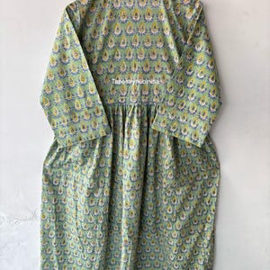 May include: A green cotton dress with a yellow and blue floral print. The dress has a round neckline and 3/4 sleeves. The dress is gathered at the waist and has a flowing skirt. The dress is made in India. The text "Tapestryhubindia" is printed on the dress.