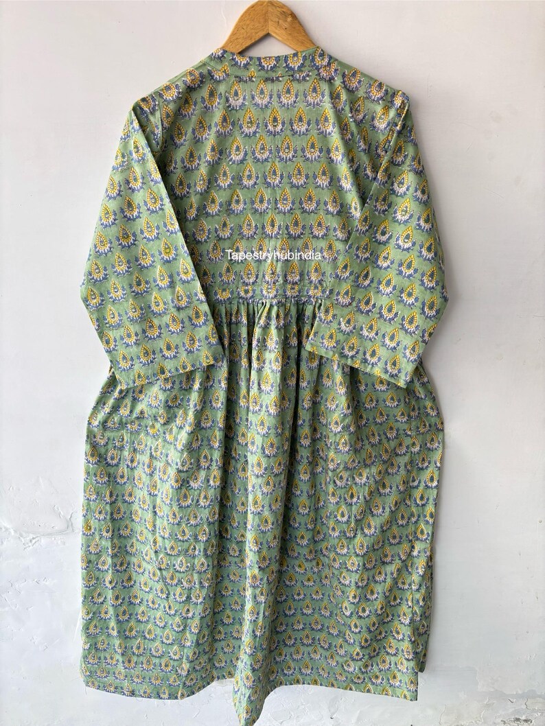 May include: A green cotton dress with a yellow and blue floral print. The dress has a round neckline and 3/4 sleeves. The dress is gathered at the waist and has a flowing skirt. The dress is made in India. The text "Tapestryhubindia" is printed on the dress.