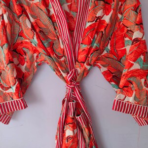 Op de afbeelding: Een rood-witte badjas met een tropische bladerenprint. De badjas heeft een strikceintuur en lange mouwen.