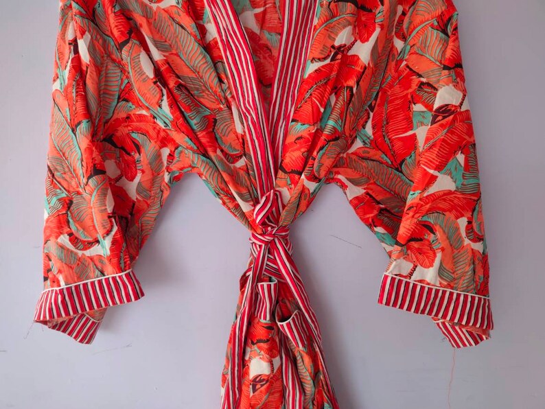 Op de afbeelding: Een rood-witte badjas met een tropische bladerenprint. De badjas heeft een strikceintuur en lange mouwen.