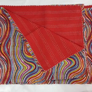 May include: A reversible quilt with a vibrant, abstract design. One side is red with white stitching, the other has a colourful, wavy pattern in red, blue, yellow, and green. The quilt is folded, showing both sides. The edges are scalloped.
