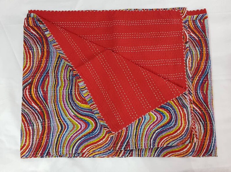 May include: A reversible quilt with a vibrant, abstract design. One side is red with white stitching, the other has a colourful, wavy pattern in red, blue, yellow, and green. The quilt is folded, showing both sides. The edges are scalloped.