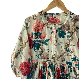 May include: A white cotton dress with a floral print featuring large pink and red flowers, green leaves, and brown branches. The dress has a button-up front and a gathered skirt.