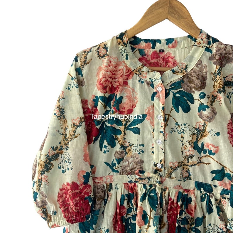 May include: A white cotton dress with a floral print featuring large pink and red flowers, green leaves, and brown branches. The dress has a button-up front and a gathered skirt.