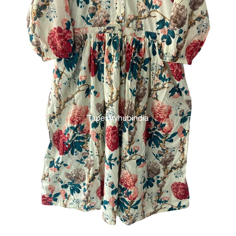 May include: A white cotton dress with a floral print featuring pink, red, and green flowers. The dress has a button-up front and a gathered skirt.