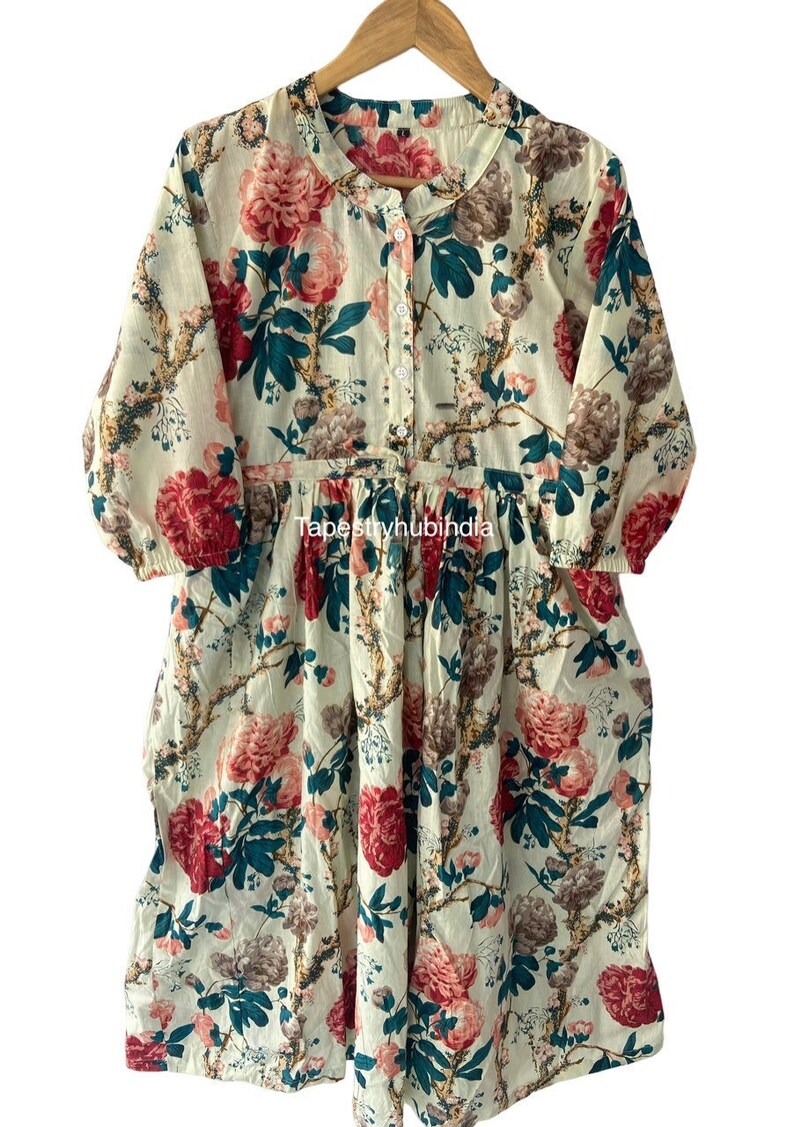 May include: A white cotton dress with a floral print featuring pink, red, and green flowers. The dress has a button-up front and a gathered waist.