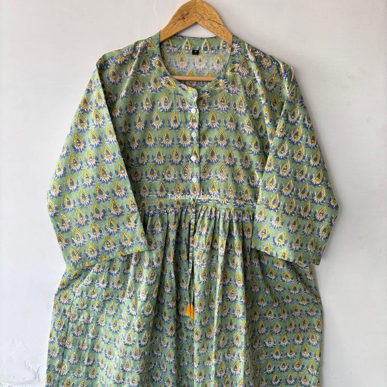 May include: A green cotton dress with a floral print and a tie at the waist. The dress has a button-up front and three-quarter sleeves.